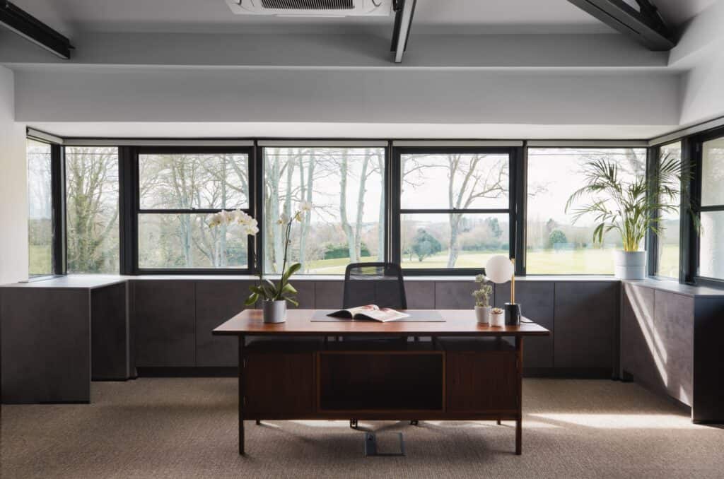 A large modern wooden desk with flowers and magazine in front of stunning winslade park views, with desk chair, lamp and modern grey cupboard fittings behind in a Winslade Park serviced office space.