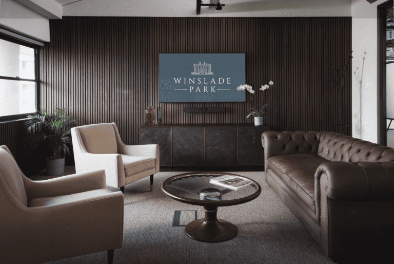 A modern serviced office with plush dark coloured leather couch, two white arm chairs, a wall-mounted screen displaying the Winslade Park logo, and coffee table in centre. Room has black paneled walls and short pile grey carpet and flowers displayed.