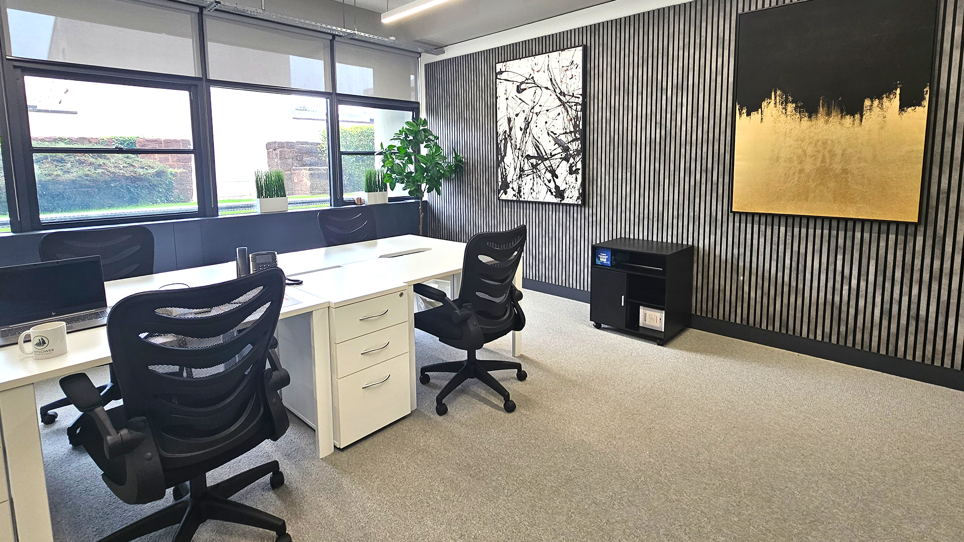 Light and airy office at Winslade Park with large windows, white desks, black ergonomic chairs, stylish grey panel walls with contemporary artwork and a tall potted plant in the corner