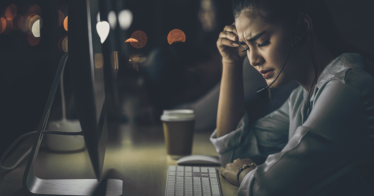 Woman appears stressed while sitting, with elbow on desk and hand on head, sullen faced, at her computer dealing with work issues late in the evening. Streetlights can be seen in the background through the window, the room is dark, a cup of coffee sits on the desk