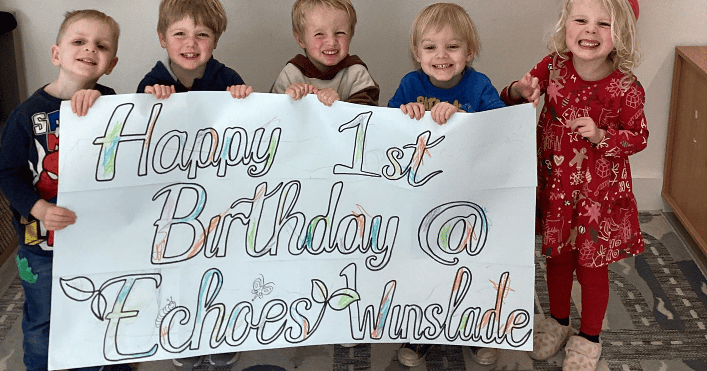 Children from Echoes Nursery hold a large happy 1 year at Winslade birthday banner up to the camera while smiling broadly
