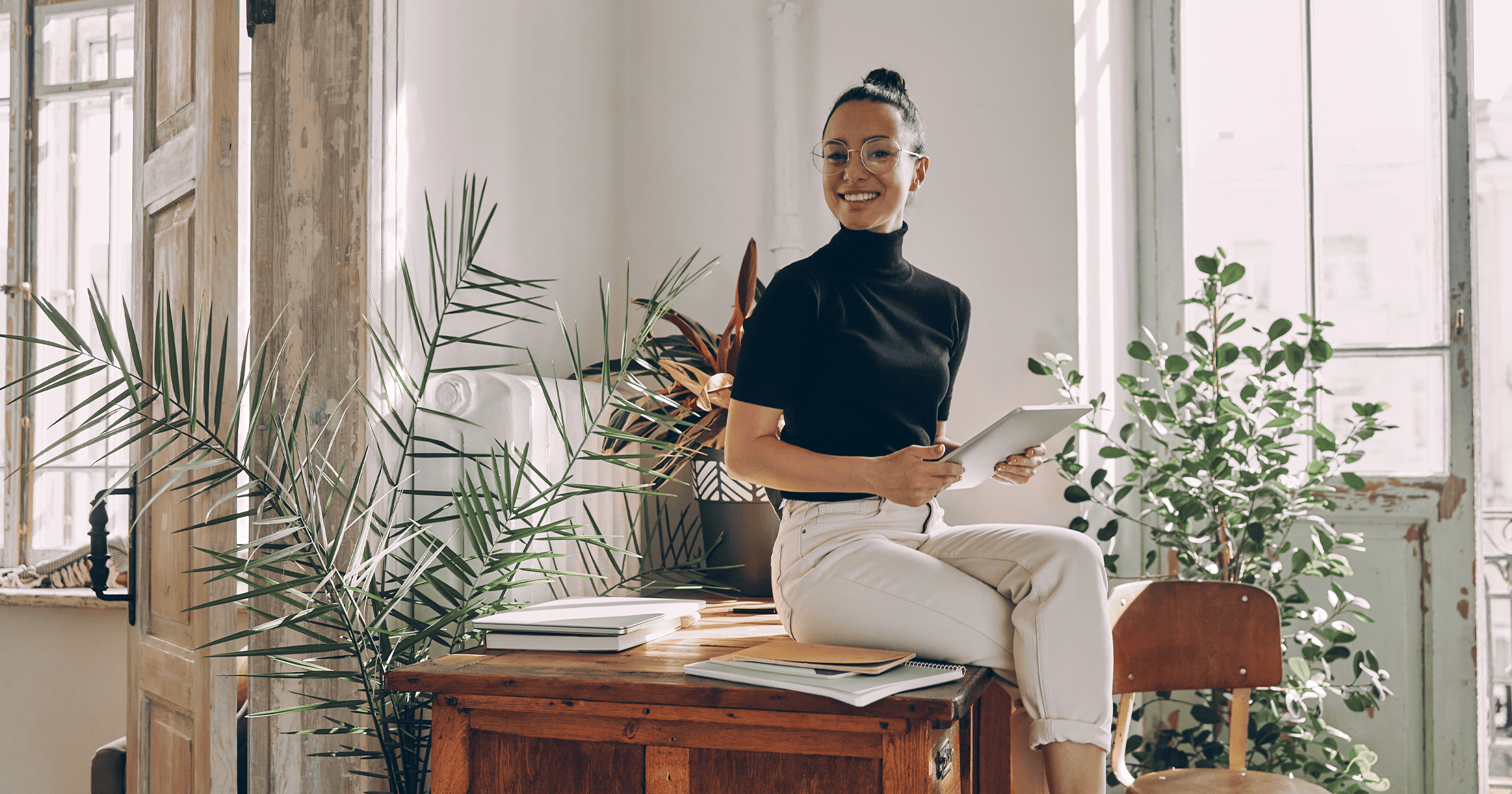 A woman sits on a desk smiling broadly surrounded by plants in an office flooded with sunlight